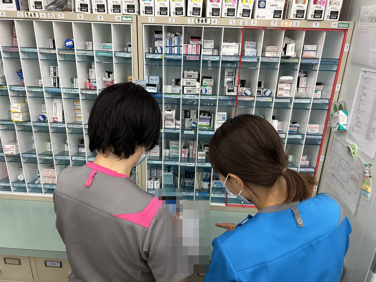 調剤棚の前で、青い服の薬剤師と一緒に薬を確認しているところ